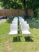 Front row view of 10 white plastic and metal chairs arranged in two columns on grass, showing seating and backrest design.