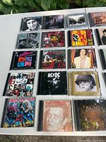 Top view of laid out 31 music CDs on white table outdoors with green leaves background, showcasing mostly 1980s rock and classic albums