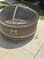 Side view of circular metal fire pit with mesh cover on top, showing rust and handle on the side