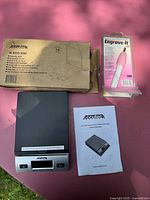 Photo showing Accuteck scale, original box, user manual, and Engrave-It engraving pen sealed in packaging on pink surface.