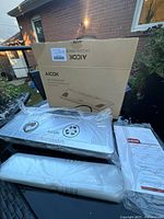 Photo of AICOK vacuum sealer, sealing bags, warranty card, and original packaging box taken outdoors on a table.