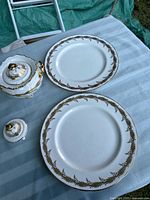 Two plates, two lids, and one handled vessel displayed on a blue cloth, showing the gold laurel and berry garland pattern and scalloped rims with gold trim