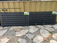 Two black plastic slotted panels leaning against a wooden fence on a paved ground area, showing full panel length and slots.