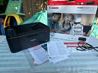 Canon PIXMA TS3420 wireless printer next to its original box, setup instructions, documents, and power cable. Printer is black with control buttons on the left side and flatbed scanner on top. Box shows branding and marketing images.