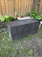 Dark grey rectangular outdoor plastic storage bin with a closed lid on grass with a wooden fence background.