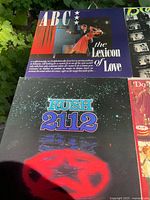 Two vinyl LP album covers on grass background featuring ABC's The Lexicon of Love with a woman and man dancing, and Rush's 2112 album with a cosmic red symbol on black background.