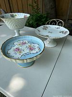 Three vintage porcelain footed serving dishes displayed on a table outdoors showing various floral patterns and shapes.