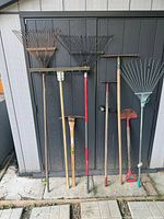 Full view of eight assorted garden tools including rakes, pick mattock, edger, scraper, positioned against a black shed door.