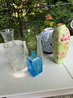 Overall view of 7 decorative vases and vessels arranged on table outdoors with garden background. Front and center is a blue square art glass bottle with teardrop appliqué, next to a tall floral hand-painted ceramic vase with yellow-green background. Visible are clear and green textured glass vases, blue and white ceramic ginger jar, and cut crystal vases.