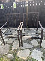 Front view of two metal brown outdoor chair frames without seating surfaces, placed on stone patio next to black fence.