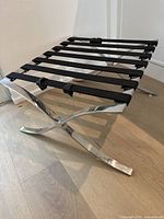 Full side view of chrome X-shaped base ottoman or luggage rack showing the black webbing strap seating surface and chrome frame on wood floor.