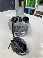 Gray Coleman electric air pump with power cord wrapped and placed on table, showing top detail with two black connectors and label with instructions on side.