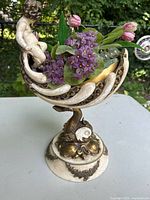 Side view of baroque-style compote showing cherub figure seated above shell-shaped bowl and dolphin pedestal base with faux purple violets and pink tulips flowers