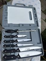 Top view of open gray hard plastic case showing six knives and sharpening steel with white cutting board inside lid.