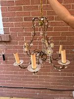 Full view of the 6-light chandelier with brass arms, cream candle sockets, and hanging chain against a brick wall.