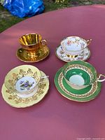 Four vintage bone china tea cups arranged on a table showing varied floral and gold designs including gold reflective, floral white, green with gold detailing, and yellow with floral center.