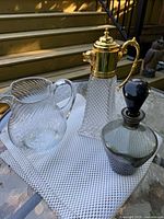 Three vintage glass pieces on a mesh table: clear ribbed Arcoroc glass pitcher, cut glass carafe with gold-tone lion-head handle and lid, smoky grey glass decanter with black stopper.
