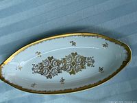 Top-down view of white elongated oval porcelain dish with central and scattered detailed gold floral designs and a heavily gilded scalloped rim.