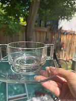 Close-up photo showing a clear glass cup with squared handles placed on a matching saucer, highlighting the circular pattern at the base and transparency of the glass under natural light outdoors.