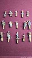 Full set of 15 hand carved wooden cork stoppers arranged on a surface, showing variety of carved figures with hats and movable parts.