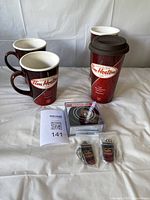 Two Tim Hortons ceramic mugs, two travel mugs, one silicone lid, two keychains and a package of four hockey puck coasters photographed on white surface.