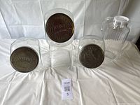 Three clear glass candy store style containers with metal screw-on lids embossed with "Moscow Candies." Also included is a clear ribbed plastic pitcher with white lid and handle.