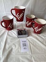 Four red ceramic Tim Hortons mugs with white interiors and recessed 'Tims' logos displayed in an upright position with packaged Senator coasters in front, all placed on a white cloth.