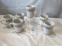 Photo showing six white ceramic coffee mugs with Tim Hortons logo arranged on white fabric background.
