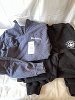 Folded men's dark gray long sleeve sweatshirt with zipper collar showing Tim Hortons logo and attached packaging tag in size 3XL, next to folded black track pants.