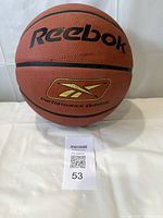 Orange Reebok basketball with black logo and gold emblem, placed on white surface with auction lot number tag.