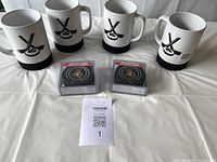 Four white mugs with black hockey puck rubber bases and two sets of packaged Ottawa Senators hockey puck coasters placed on a white cloth background.