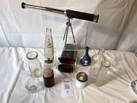 Photo showing vintage Tasco telescope on tripod, bottles, jars, small vases and holders arranged on table.