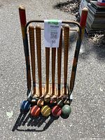 Photo showing six wooden croquet bats placed upright in rusty metal wheeled carrier with matching colored balls on asphalt ground in sunlight.