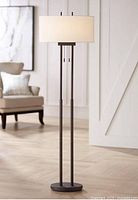 Front view of the lamp in a staged setting showing the bronze metal base, dual rods, and off-white drum shade.