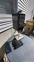Full view of the black and silver Rockwell drill press on a round surface outside. Shows drill head, column, and base.