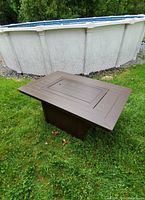 Rectangular outdoor fire pit table on green grass near above-ground pool. Bronze/brown metal table frame with recessed center panel and base pedestal.