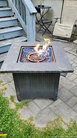 Fire pit table lit up with visible flame over blue glass rocks in square burner, dark gray stone-like top, wicker-style black base, set on paved surface next to steps and outdoor chairs.