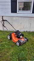 Side view of orange and gray Husqvarna lawnmower on grass showing body, engine, wheels, and partial handle