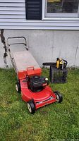 Photo of Toro red lawnmower with beige rear bag on grass and handheld yellow-black chainsaw on crate beside it.