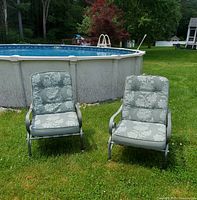 Two gray metal outdoor reclining chairs with green and white floral pattern cushions set on grass near a pool.