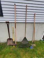 Full view of five outdoor tools leaning against a wall, including a shovel, mattock, small rake, large leaf rake, and hoe.