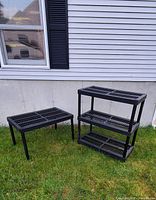 Two black plastic shelving units placed outside on grass. One has three shelves, the other has two shelves distinct. Shows overall condition and construction style.