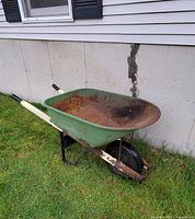 Full view of Scott's green painted metal wheelbarrow with rusted tub and frame, single black rubber tire, white handles with black grips standing on grass against a concrete and siding wall.