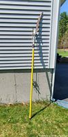 Full view of yellow pole saw leaning against wall on grass with long curved serrated saw blade and pulley mechanism.