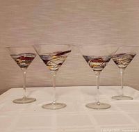 Four vintage martini glasses with colorful stained glass style painted decoration on bowl, shown side by side on white surface in front of beige background.