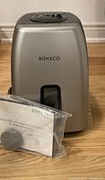 Front view of silver BONECO humidifier with user manual and replacement part sealed in plastic bag in front.