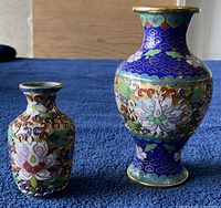 Two cloisonné vases side by side showing size difference and designs on deep blue backgrounds with gold accents.