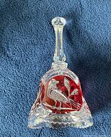 Clear crystal bell resting on a dark blue textured surface showing the bird and floral etching with red accents.