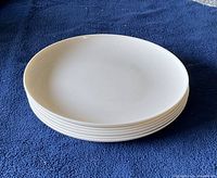 Stack of six white round ceramic dinner plates with slightly raised edges on dark blue fabric surface.
