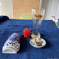 Four items displayed on blue cloth including Hard Rock Cafe glass mug, red bell pepper figurine, decorative plate, and wall pocket vase.